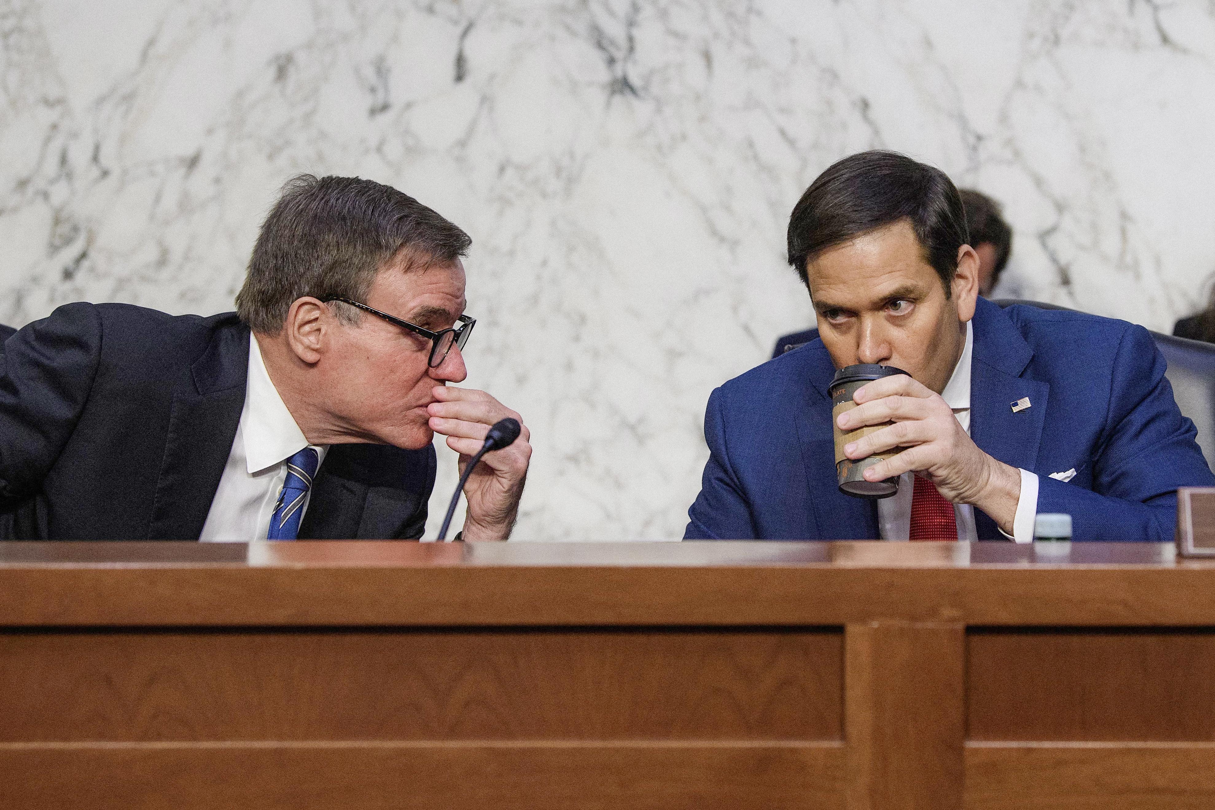 Warner leans over to Rubio to tell him something, pinching around his mouth, as Rubio sips from a paper coffee cup.