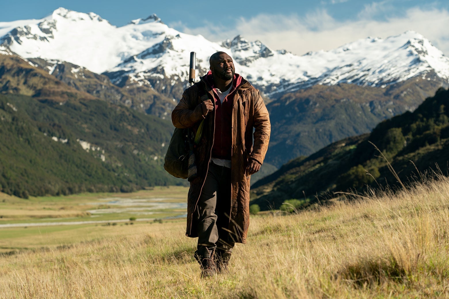 Nonso Anonzie walks across a golden meadow, with whitecapped mountains in the background. He is wearing a long leather coat and has a rifle strapped to his back. 