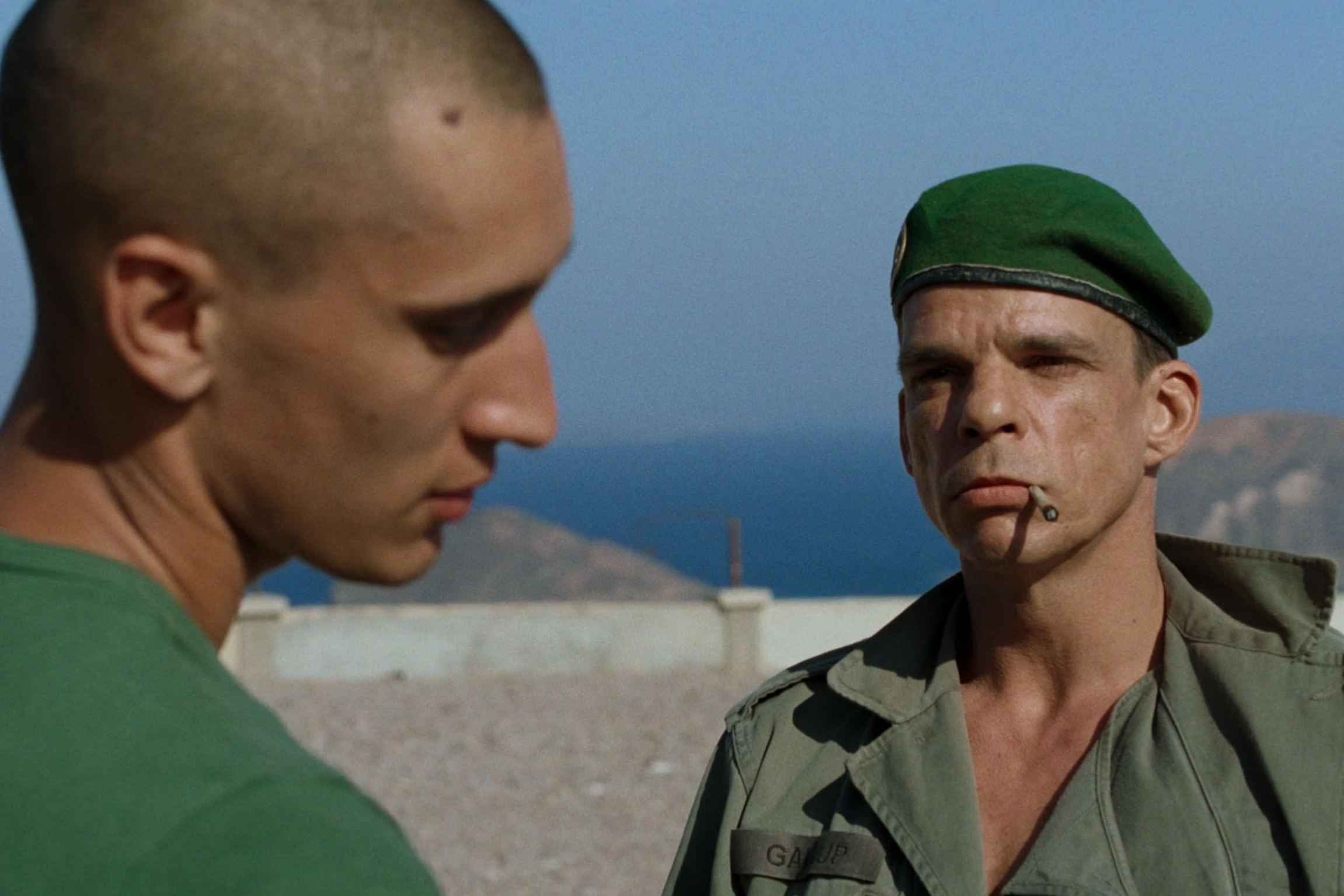 A young soldier with his back to the camera looks away while his chief warrant officer stares at him through squinted eyes with a cigarette hanging off his lips.