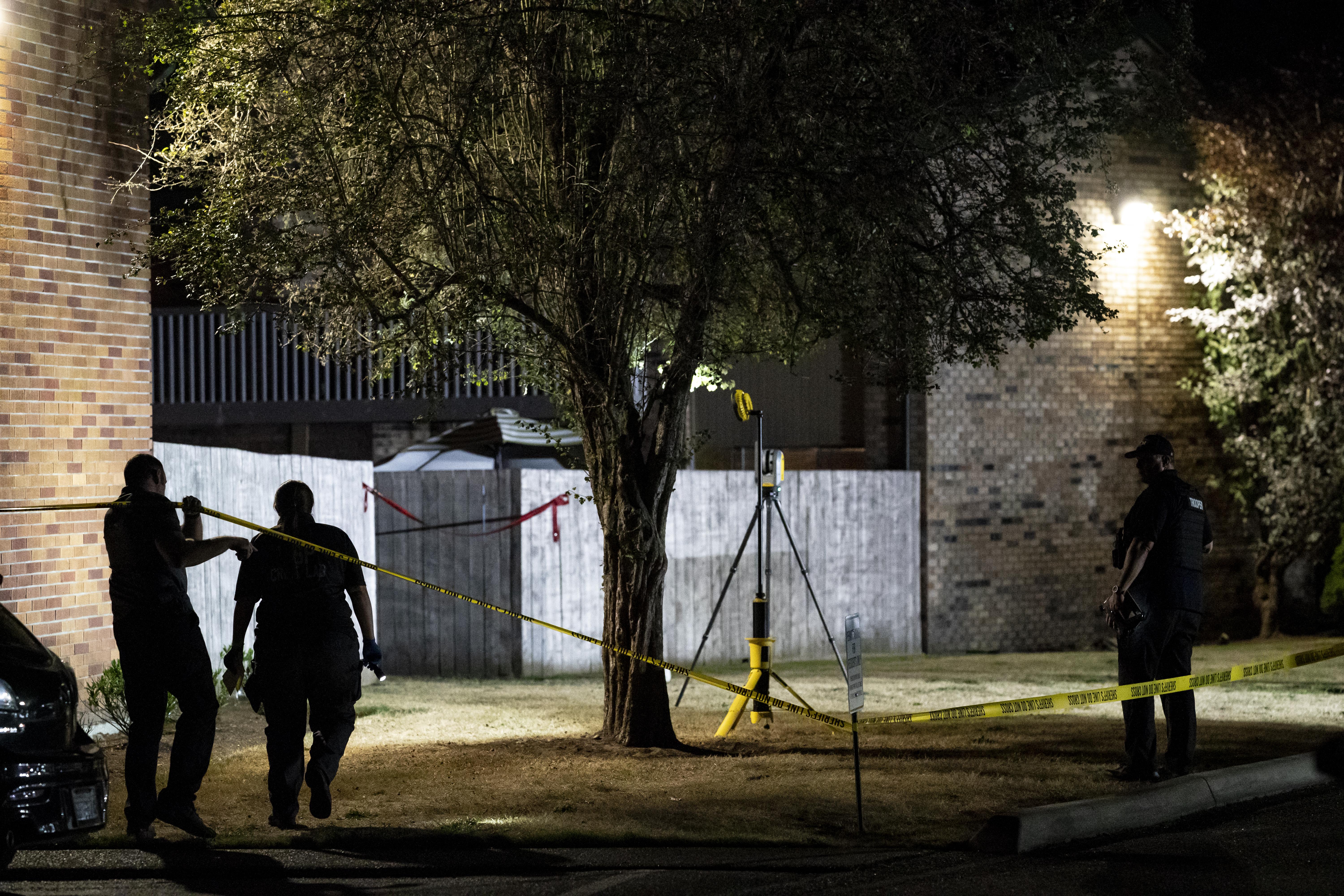 Three officers walk around a crime scene, marked by yellow tape. The scene appears to be outside an apartment building and around a tree.