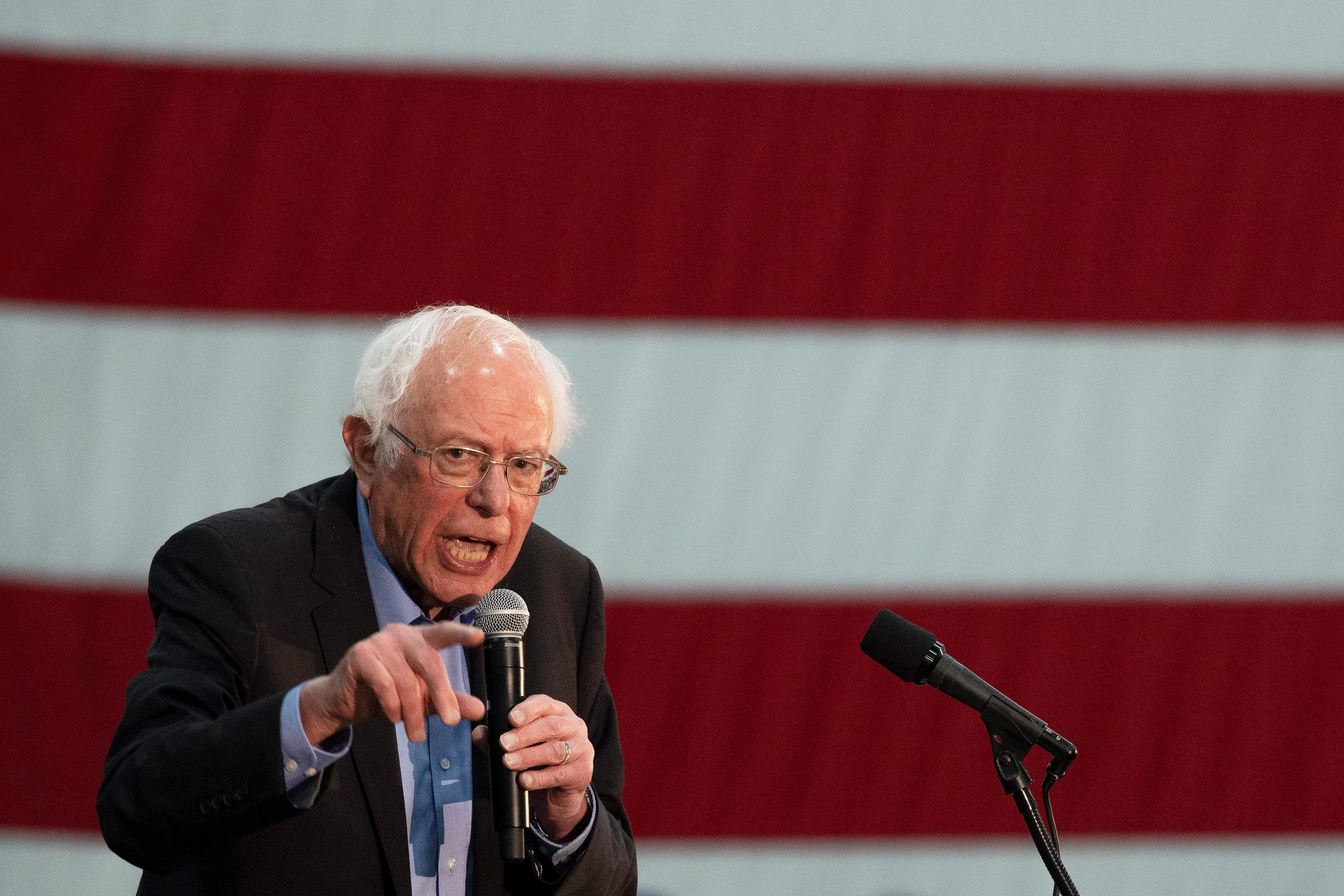 Bernie Sanders, standing in front of an American flag, speaks into a microphone.