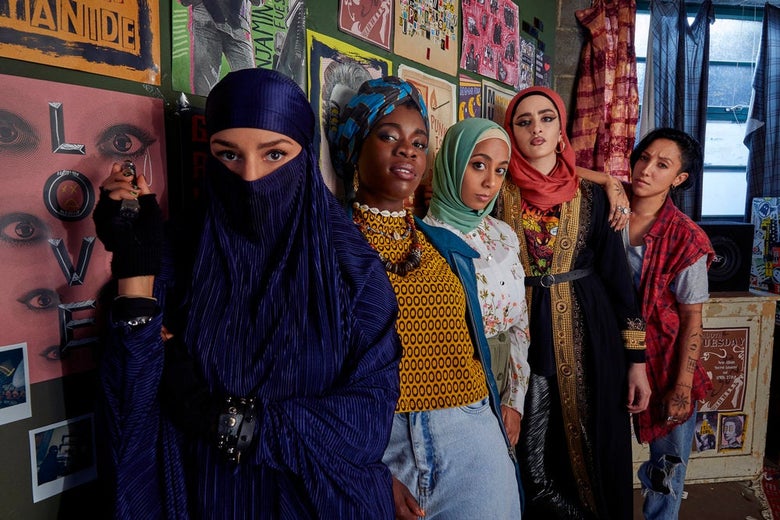 Five women lined up against a wall covered with posters of punk bands. Three of the women wear headscarves, one a niqab. They are all mugging for the camera like rock stars.