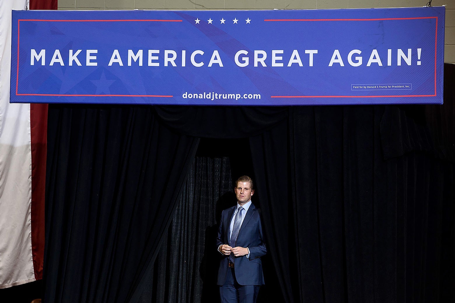 Eric Trump stands in the wings of a stage with a "Make America Great Again" sign over him.