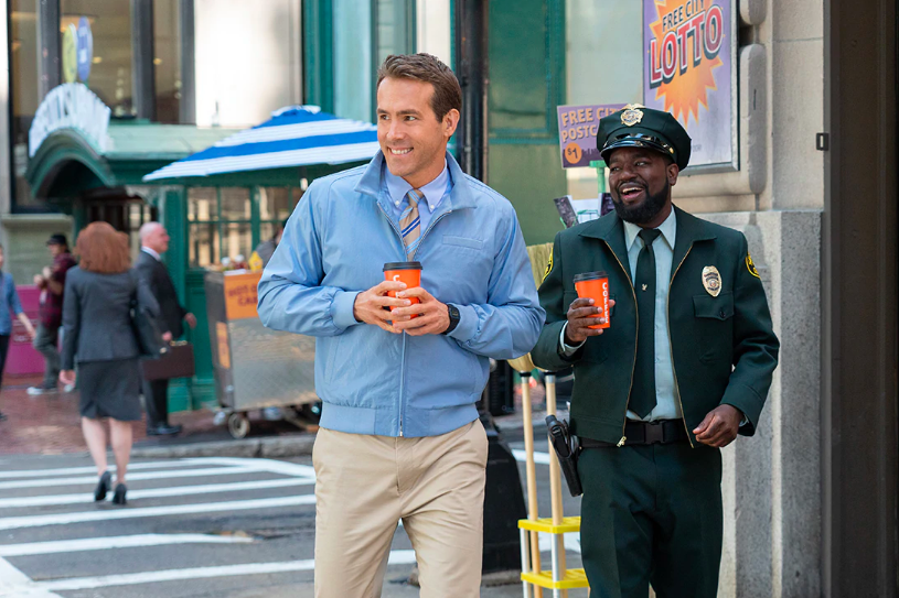 Ryan Reynolds and Lil Rey Howery stand smiling and holding to-go cups on a city street.