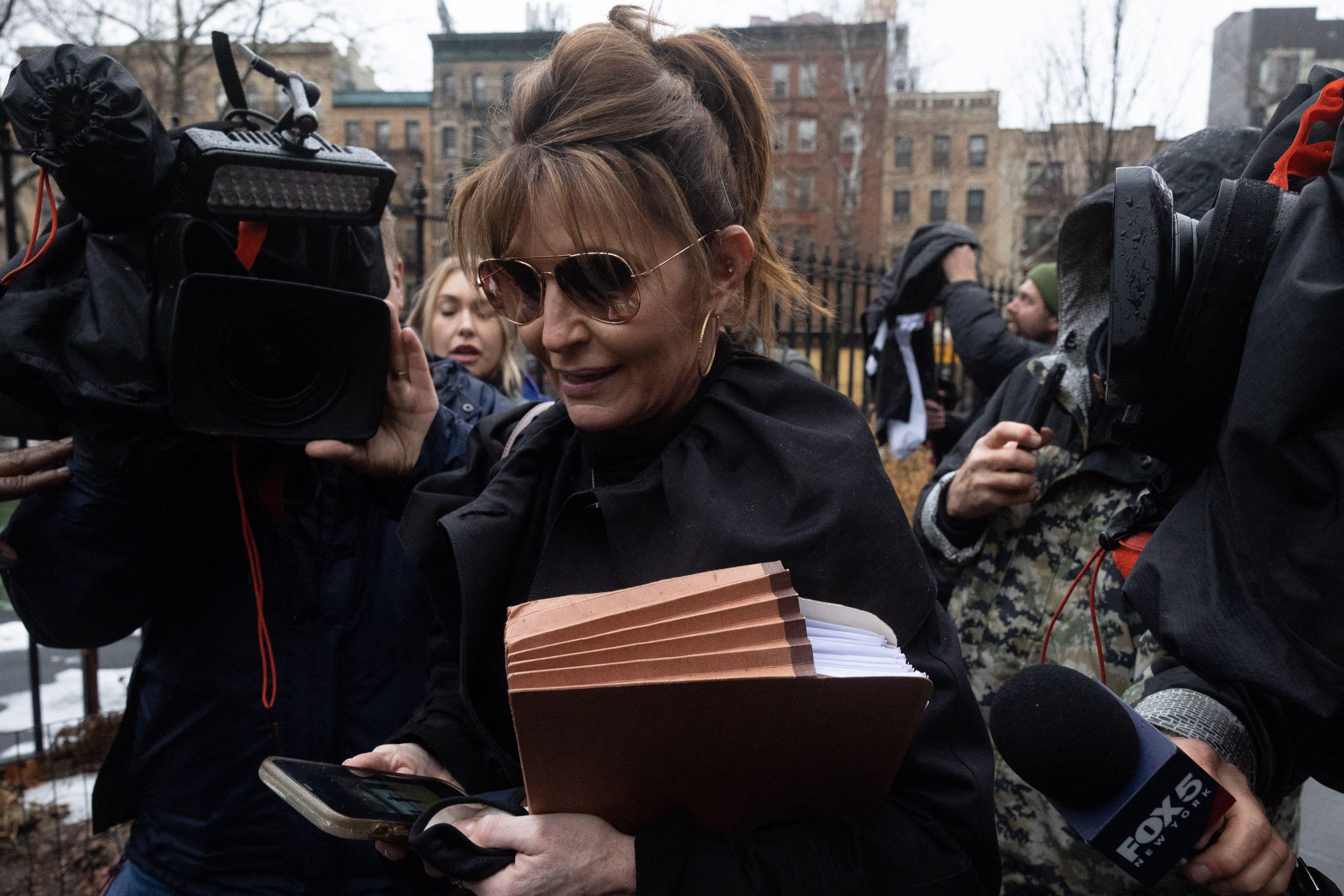 Members of the press surround Palin as she walks outside looking at her phone and holding an accordion file in the crook of her arm