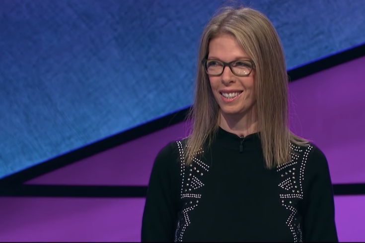 Jackie Fuchs stands in front of a blue and purple Jeopardy! backdrop, smiling in glasses and a black shirt.