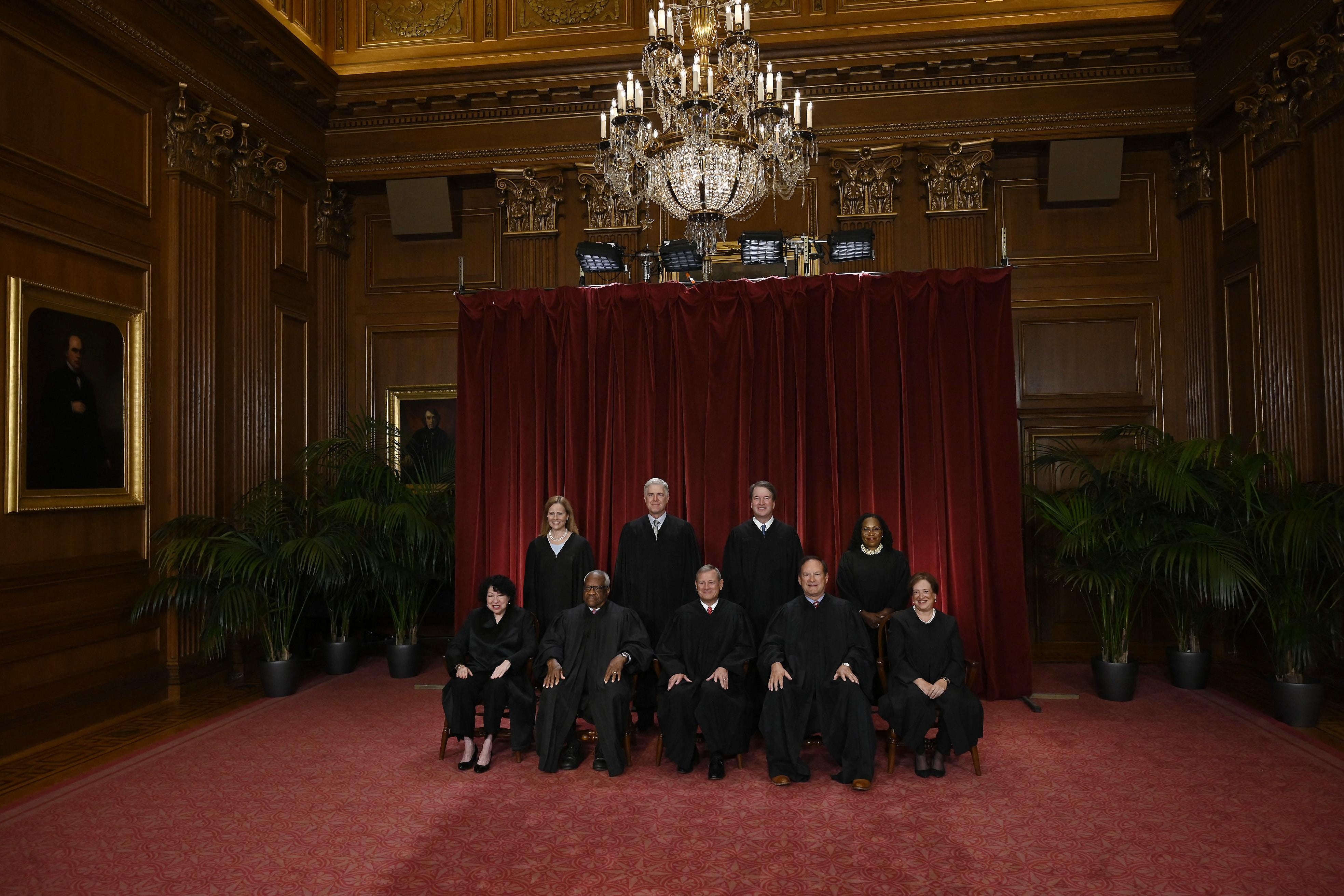 The Supreme Court poses for its official photo. This photo is further back, and you can see the justices in front of a red curtain, with the fancy room of the court surrounding them.