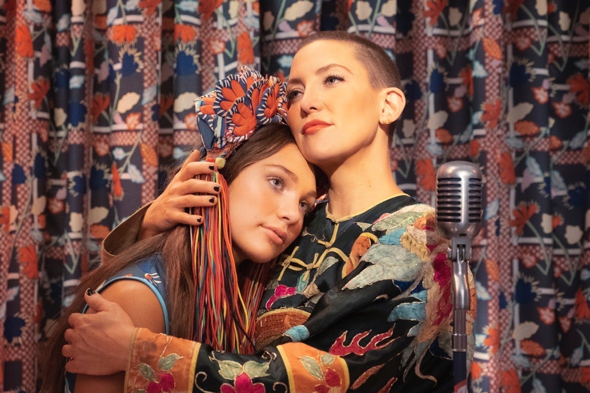 In front of a patterned curtain, Kate Hudson, with close-buzzed hair, embraces Maddie Ziegler. A microphone stands next to them.