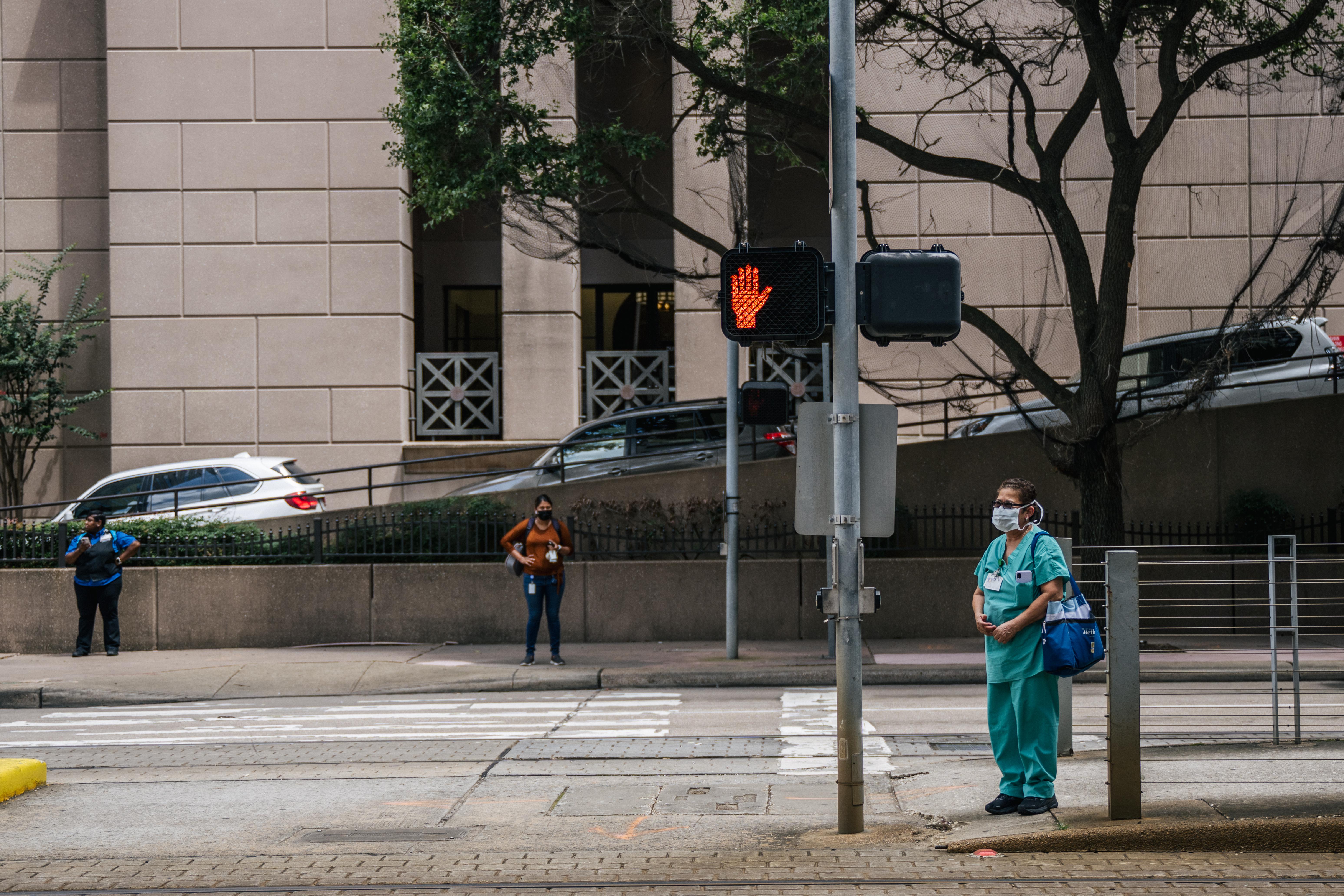 A nurse and pedestrians wait to cross an intersection outside of the Houston Methodist Hospital on June 09, 2021 in Houston, Texas. Houston Methodist Hospital has suspended 178 employees without pay for 14 days for their refusal to comply with its COVID-19 vaccine requirement. 