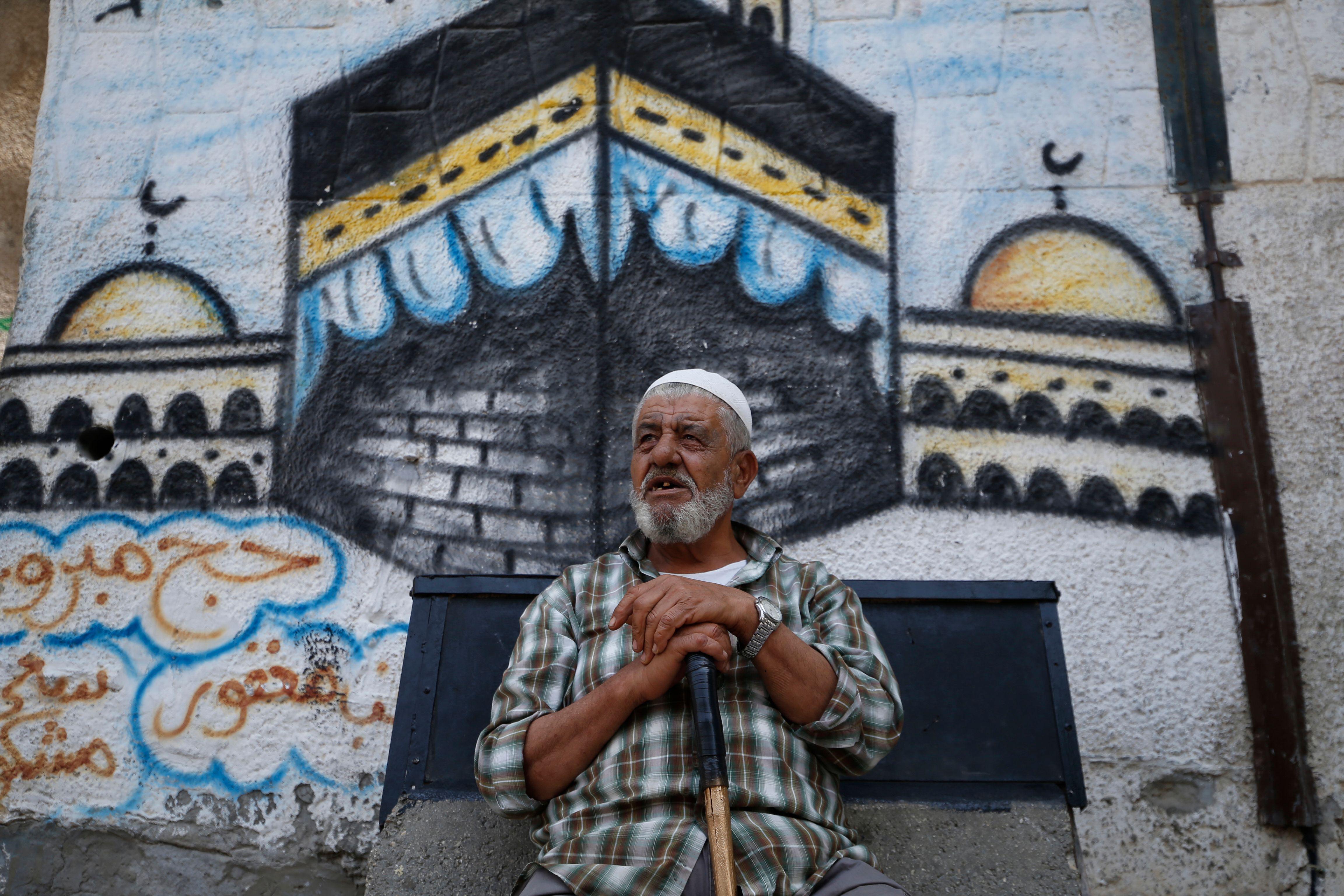 An elderly Palestinian man sits in front of a mural depicting the Kaaba.