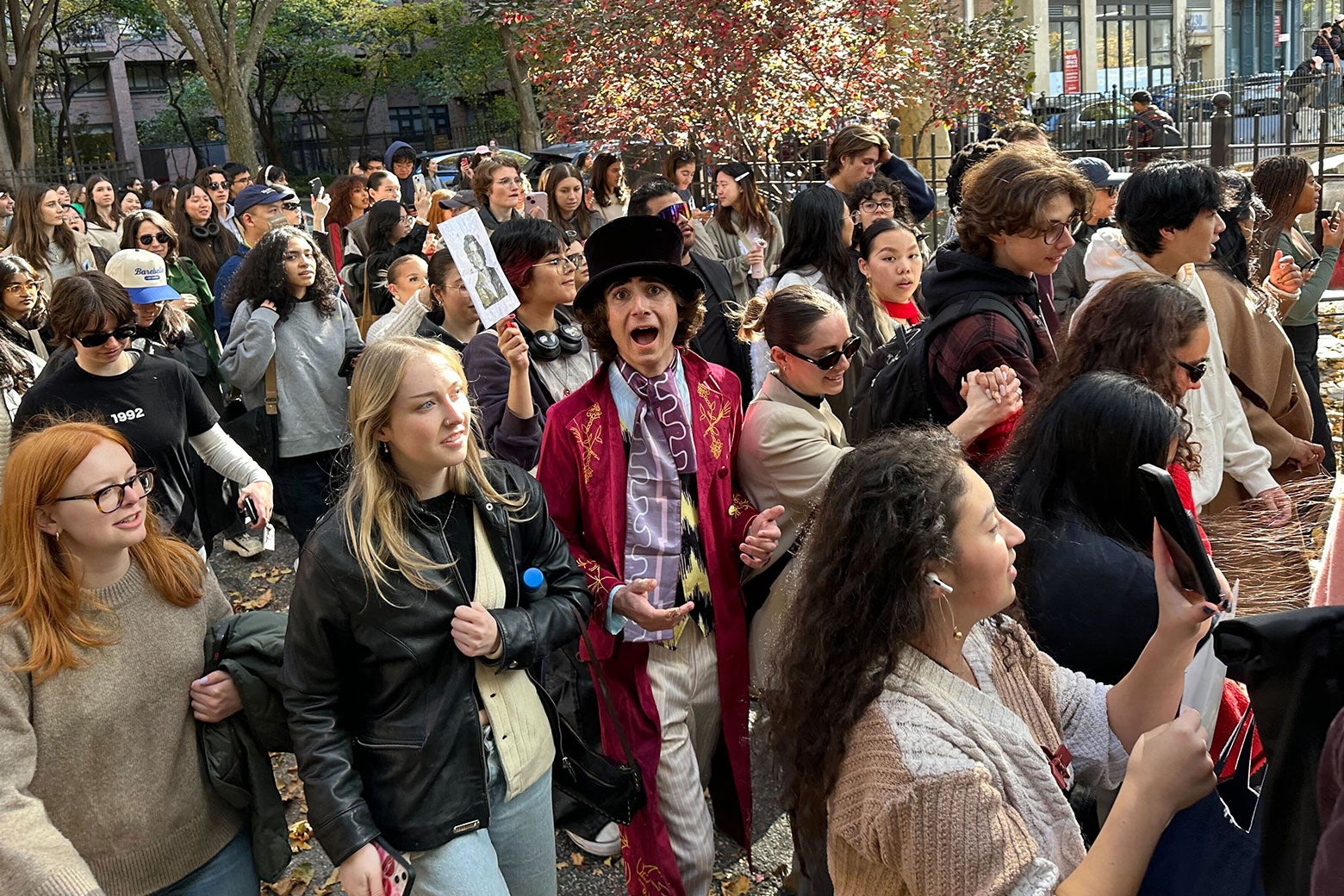 A young man resembling Timothée Chalamet, dressed as Willy Wonka, walks among a crowd of people. While everyone is looking ahead, he looks animatedly at the camera with his mouth open and eyes wide. 