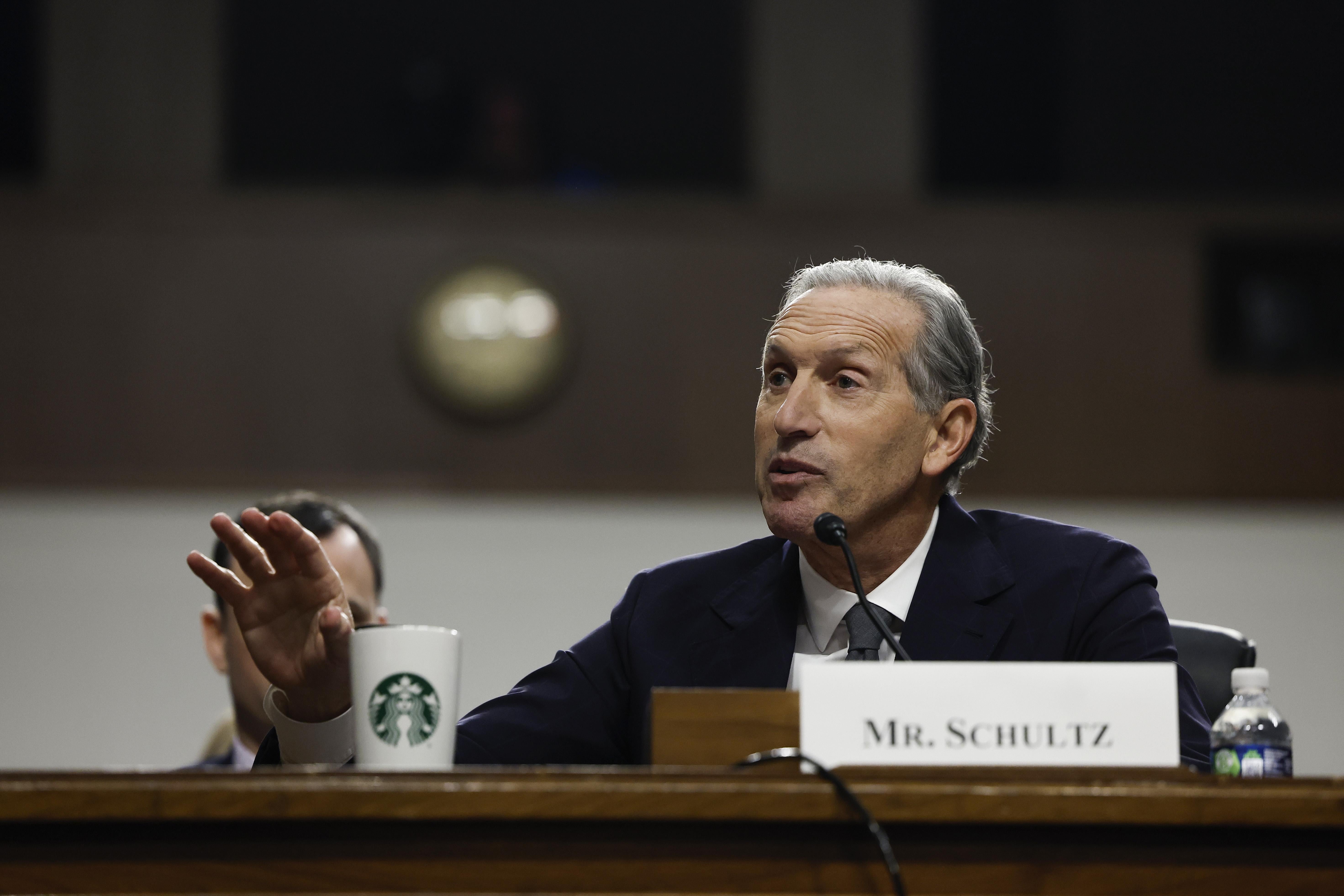 A white man with gray hair, wearing a blue suit, sits at a desk with a Starbucks branded cup next to him. A white placard in front of him reads "Mr. Schultz."