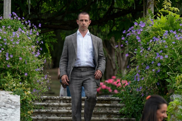 A man in a gray suit with his brow furrowed walks down steps in a garden