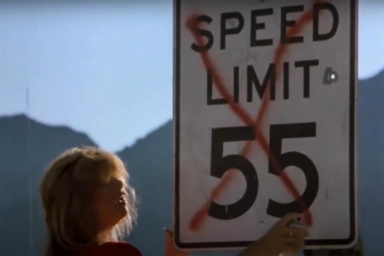 A woman uses red spray paint to put an X over a SPEED LIMIT 55 sign. 