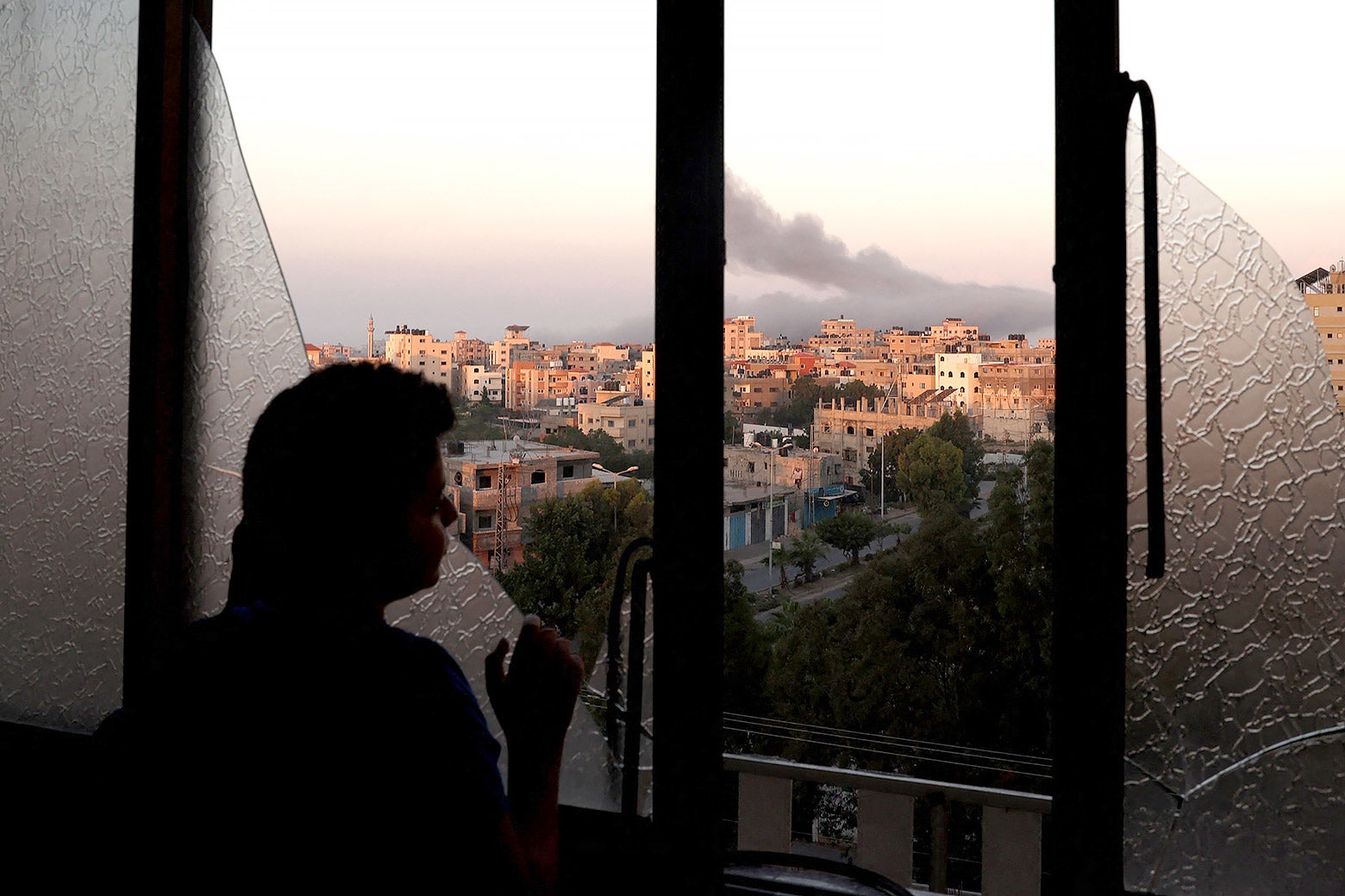 A child looks out a broken window at smoke billowing behind the buildings in the distance.