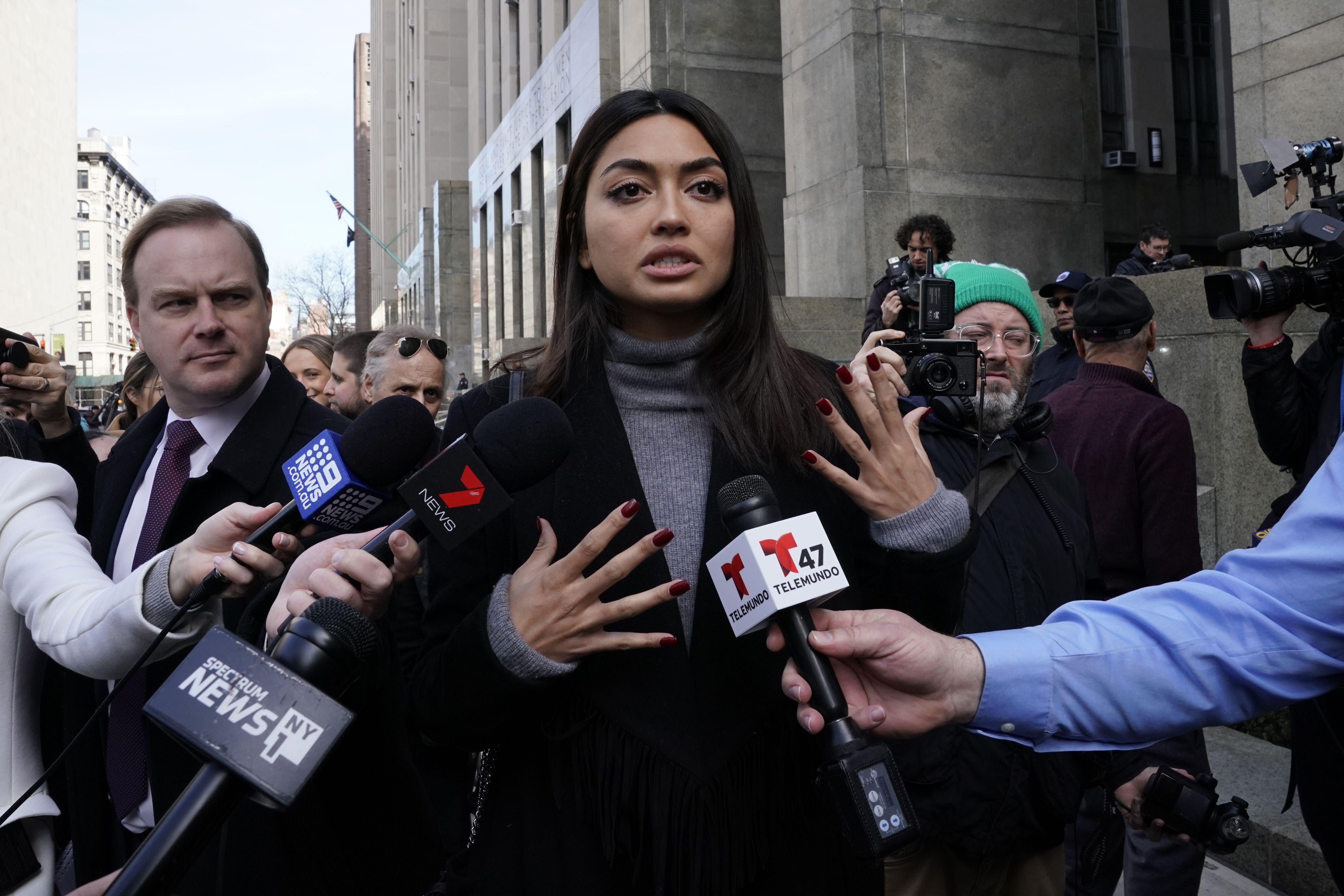 A woman in a black jacket and turtleneck stands outside a courthouse talking to reporters, surrounded by microphones. 