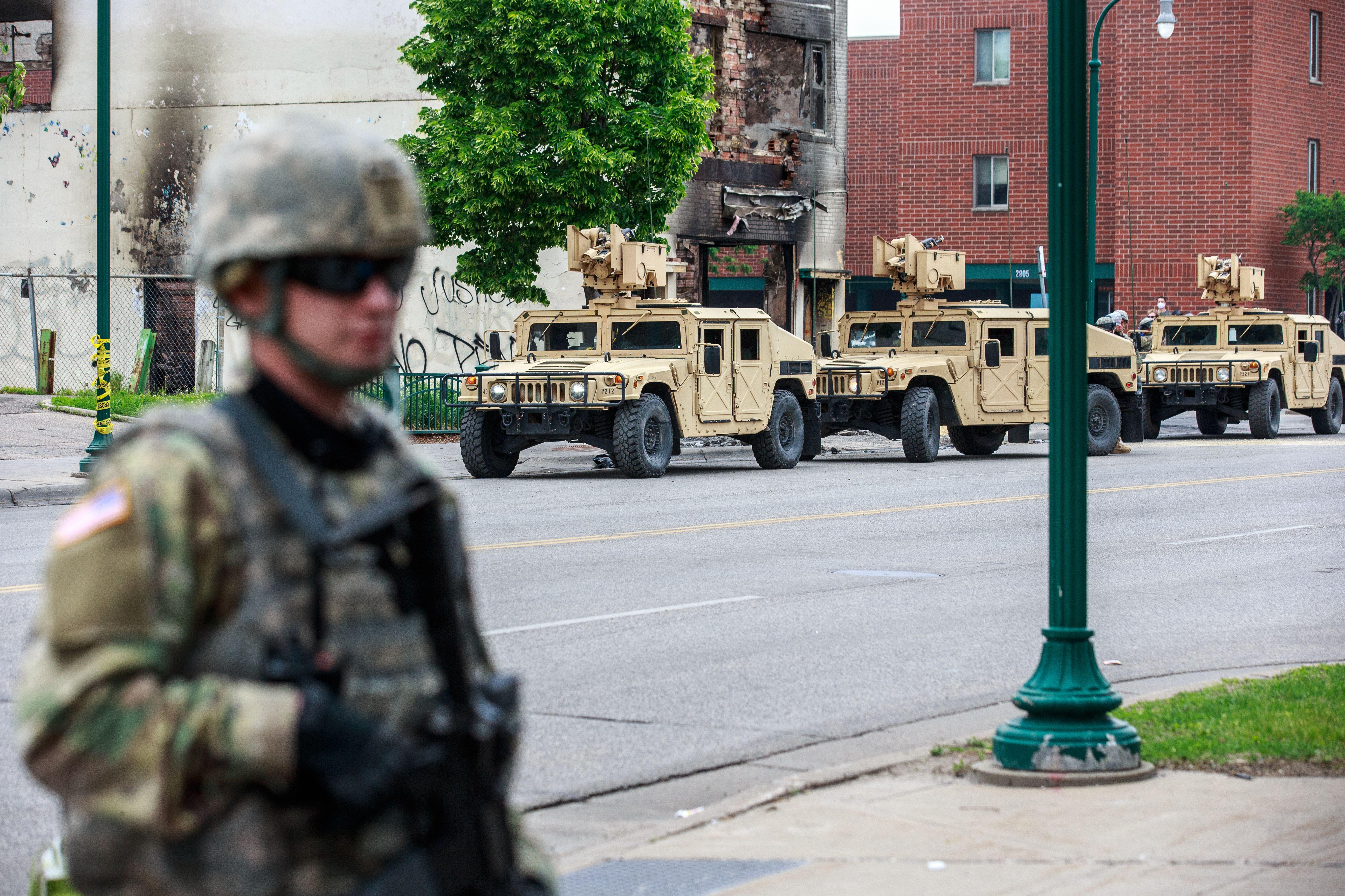 Armored vehicles roll down the streets of Minneapolis. A uniformed guardsman stands in the foreground.