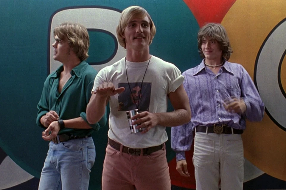 Three guys, dressed in 1970s style clothing, stand in front of a colorful building facade. All three looking in different directions. In the middle is Matthew McConaughey, who is holding a drink. 
