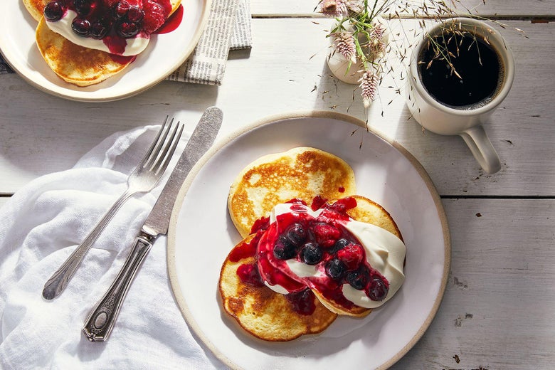 A stack of three tiny pancakes topped with fruit compote and dollops of cream. On the table nearby, a mug of dark liquid and a vase of dainty flowers.