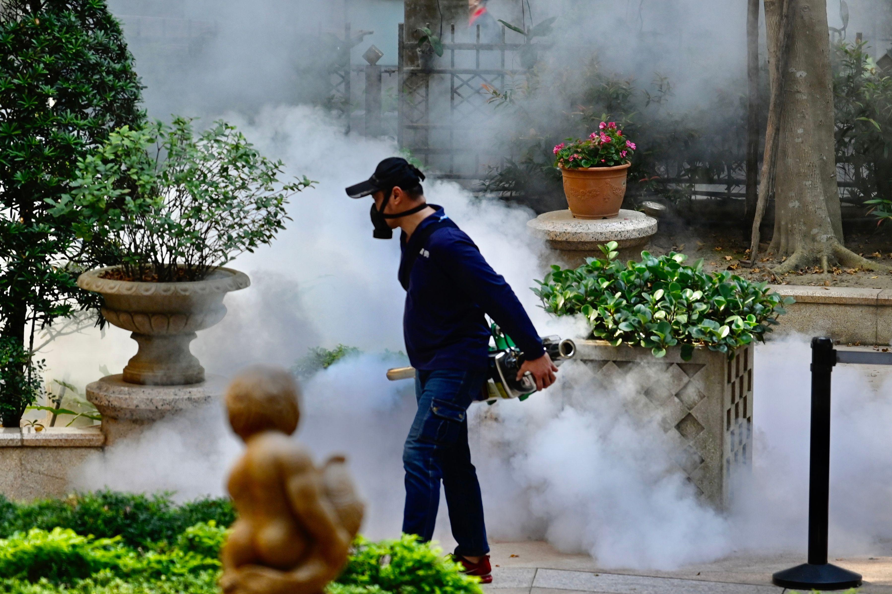 A mask-clad worker with a handheld machine walks on a street amid potted plants before billows of smoke. 