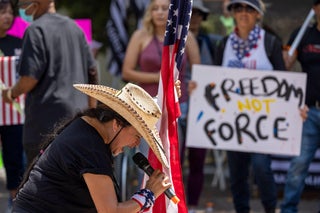 A woman in a cowboy hat and leans forward toward a microphone she's holding. With the other hand, she holds an American flag. In the background, a person holds a sign saying 