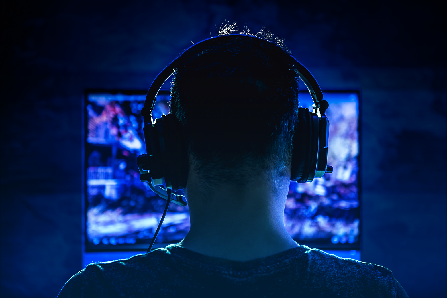 Video gamer wearing a headset sits in front of a computer screen in the dark.