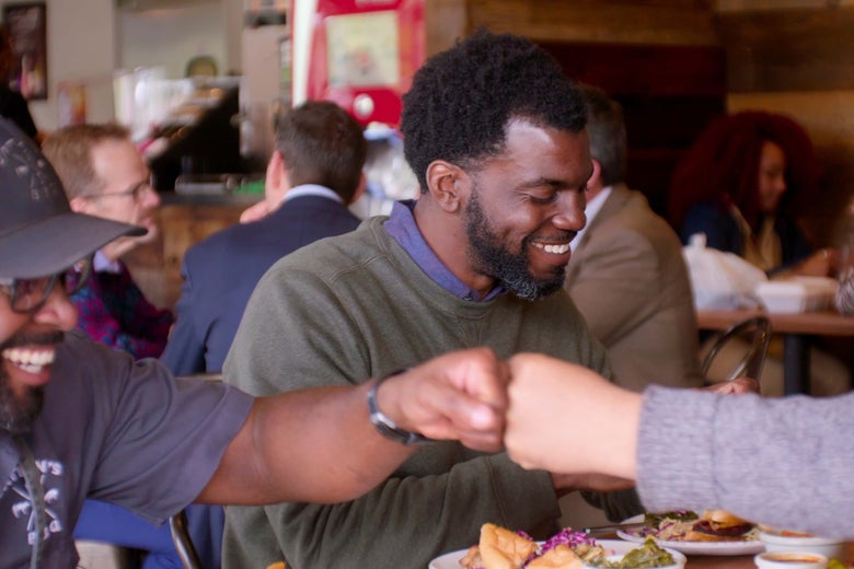 One Black man smiles as another Black man fist bumps across a table laden with food