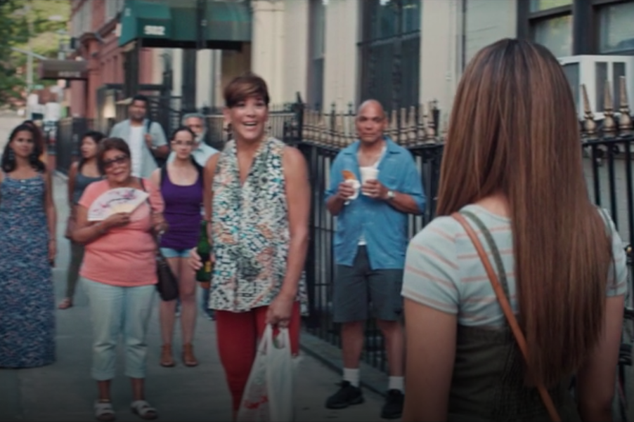 Doreen Montalvo stands amidst a crowd of people on a sidewalk. In the foreground, Nina stands with her back turned to the camera.