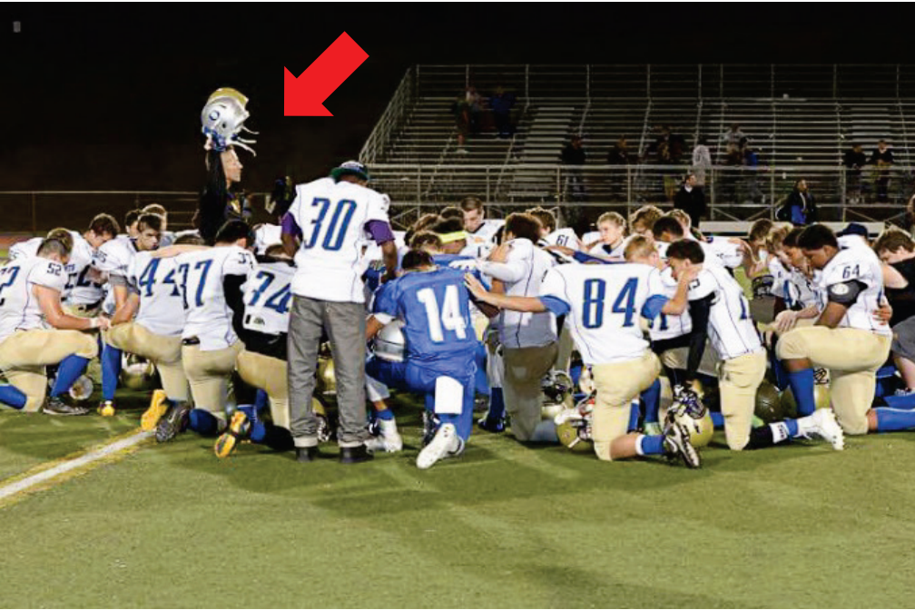 Coach Kennedy leads a prayer circle on the field, holding up a helmet. 