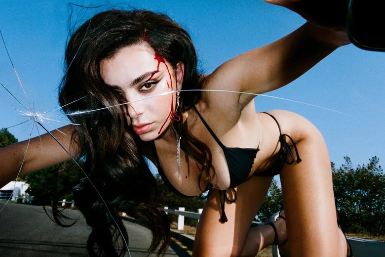 Charli with a bleeding head wound and wearing a black bikini leans over a cracked car windshield