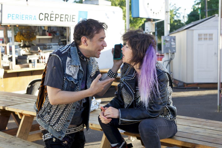 Brownstein and Armisen, in character as punks (with stereotypical jean jackets, studded accessories, and colored hair) argue into a cell phone.