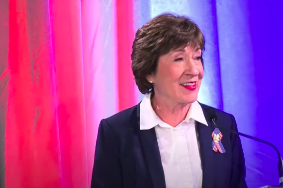 Susan Collins, in a blue suit jacket, white shirt, and American flag lapel ribbon, smiles behind a microphone against a backdrop of red, white, and blue curtains.