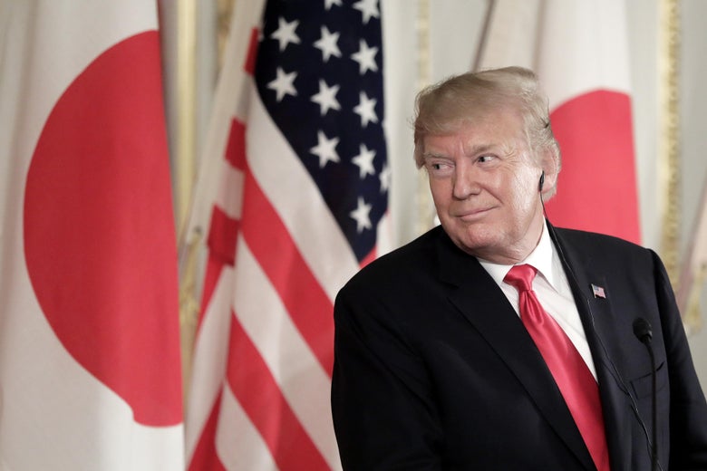 Trump smirks at a podium, with the American flag and two Japanese flags behind him.