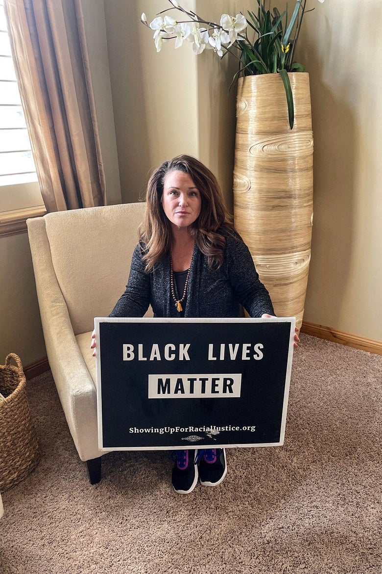 A woman sits in a chair, near a window and a plant, while holding a Black Lives Matter lawn sign.