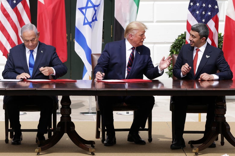 Trump speaks to Nahyan while Netanyahu signs the papers in front of him. The three are seated at a desk in a row, in front of their countries' flags.