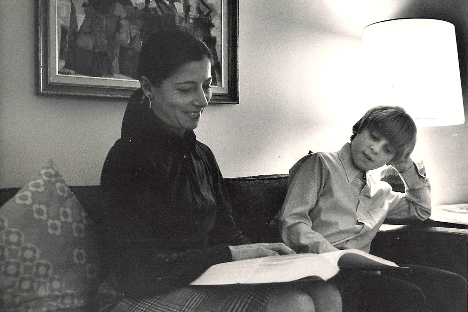 A black-and-white photo in which RBG sits on the couch with a book open next to her young son.