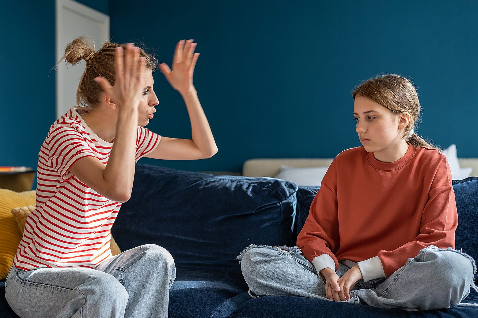 An upset-looking mother sits on a couch and talks to her daughter, who is sitting quietly and looking away.