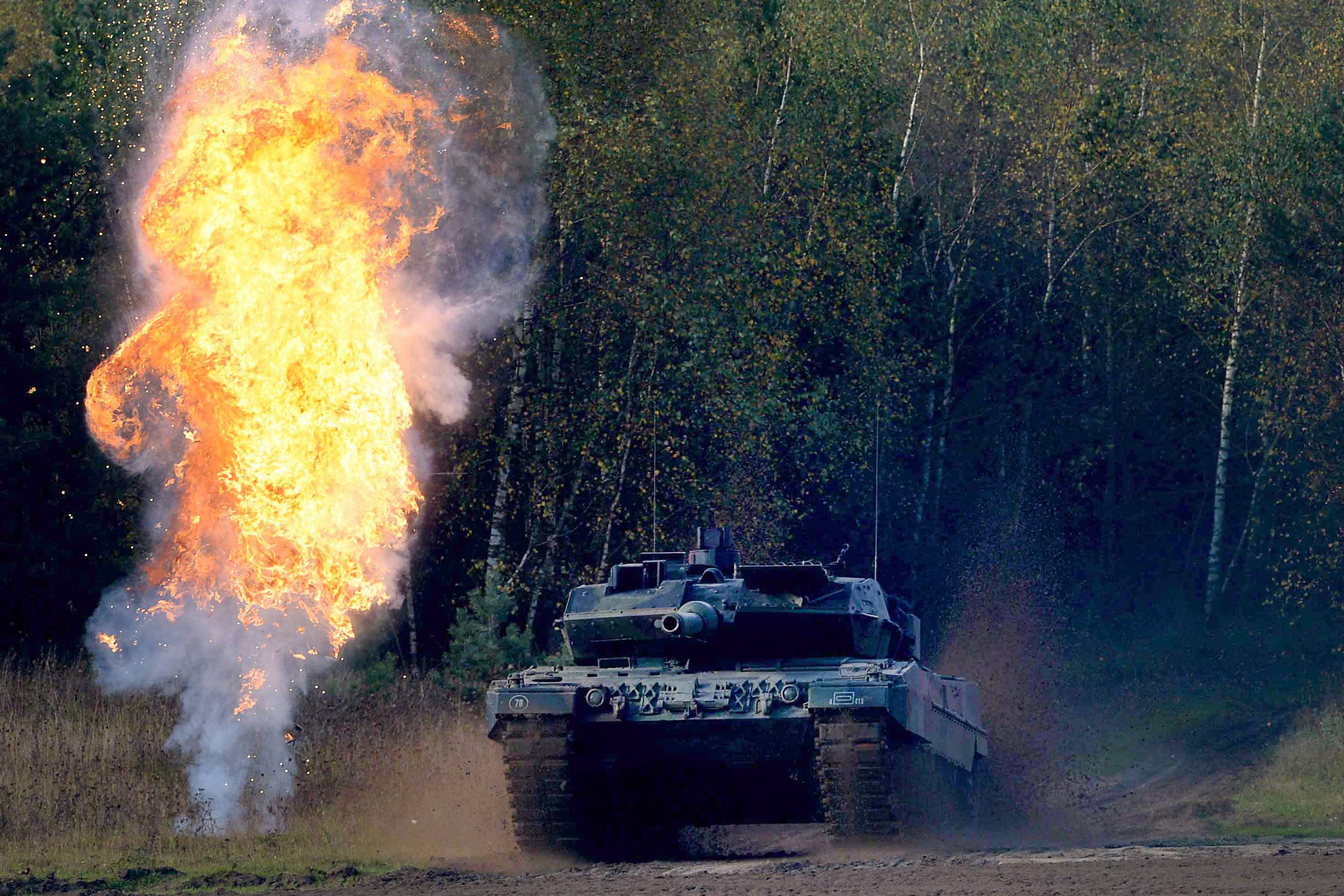 A big tanks rolls across a field with fire beside it.