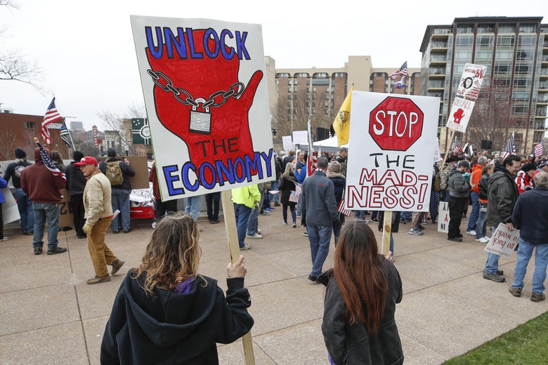A crowd of protesters hold signs with slogans such as "Unlock the Economy" and "Stop the Madness"