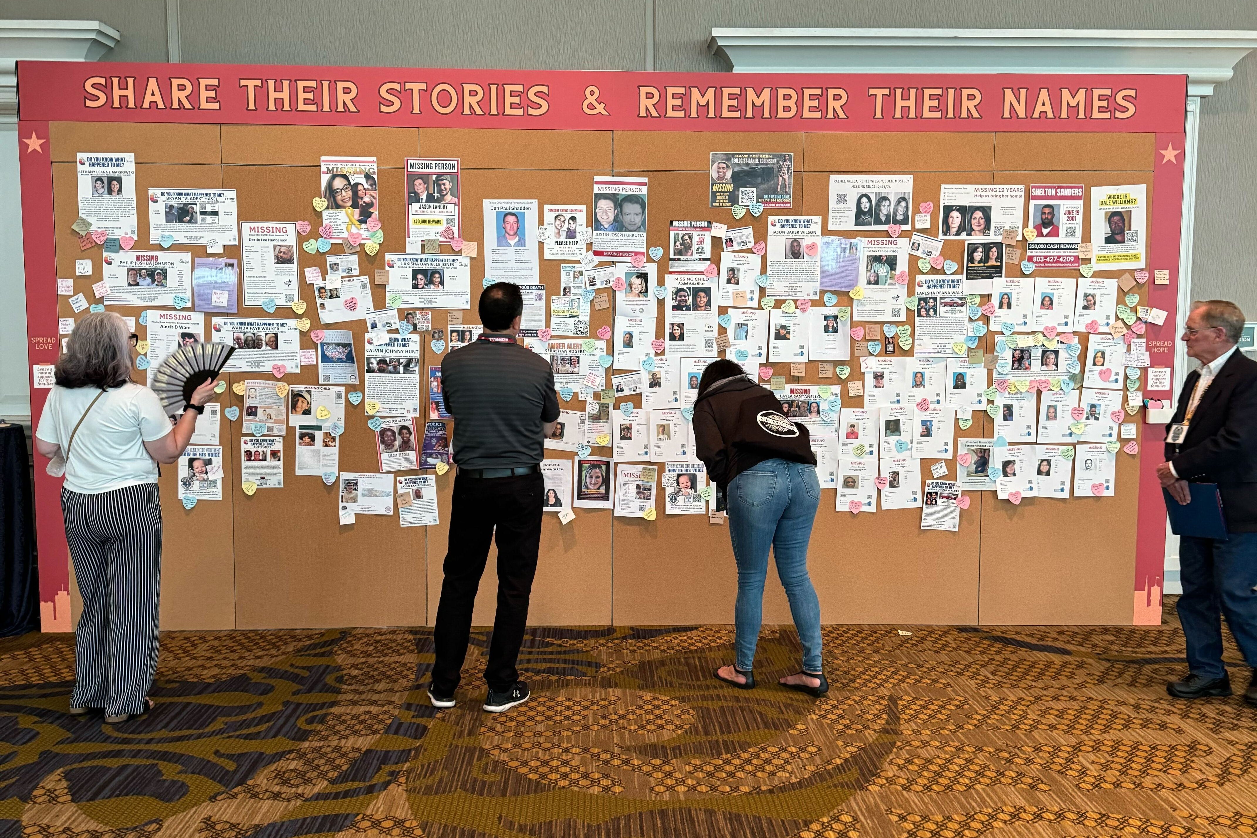 A large corkboard features a banner that says, "Share Their Stories & Remember Their Names." Underneath are photos and bios of the deceased. Multiple passersby have stopped to look and read the stories.