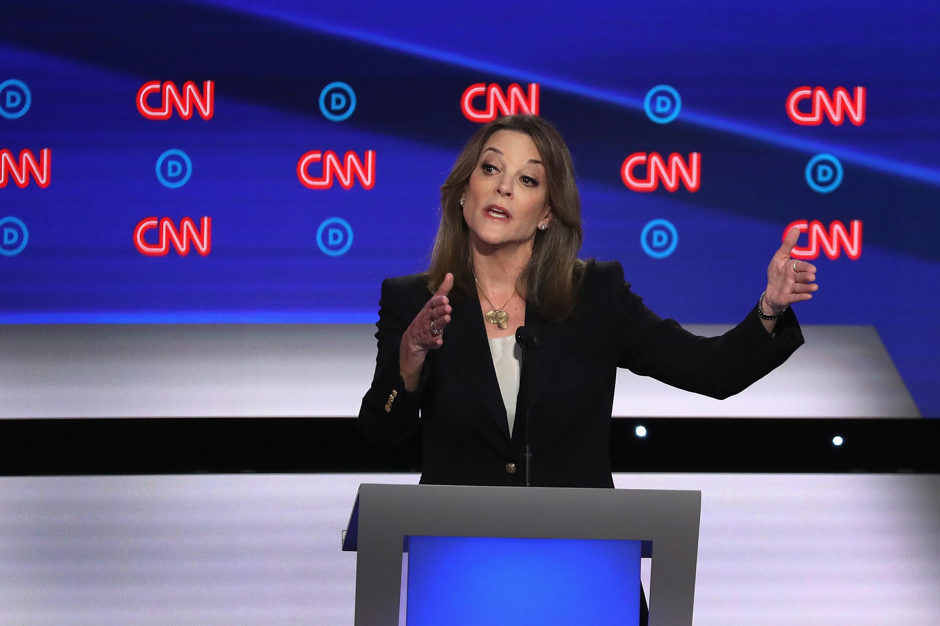 Marianne Williamson, in a dark suit, stands against a debate backdrop with the CNN logo repeating on it and makes a gesture with her hands as if measuring something in the air. 