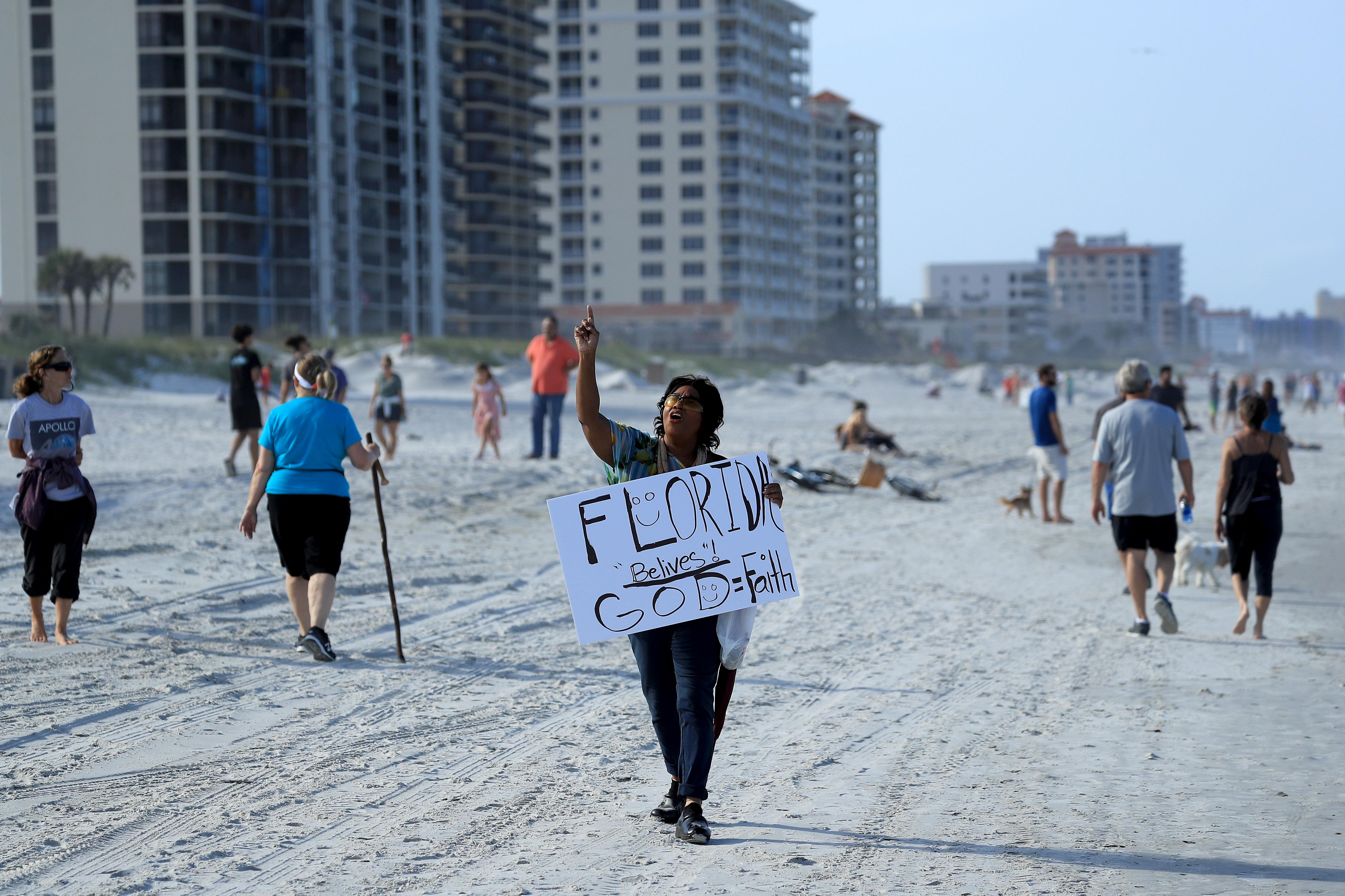 People walking on the beach. One person carries a sign that says "Florida belives! God = Faith."