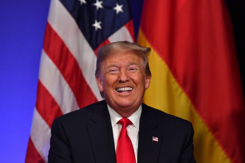 Trump smiles while sitting in front of an American and German flag.