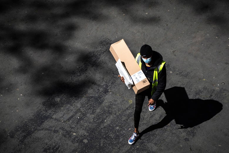 An aerial view of a person walking alone on a street. They're wearing black clothes, blue shoes, and a surgical mask, and they're carrying a box and a small parcel.