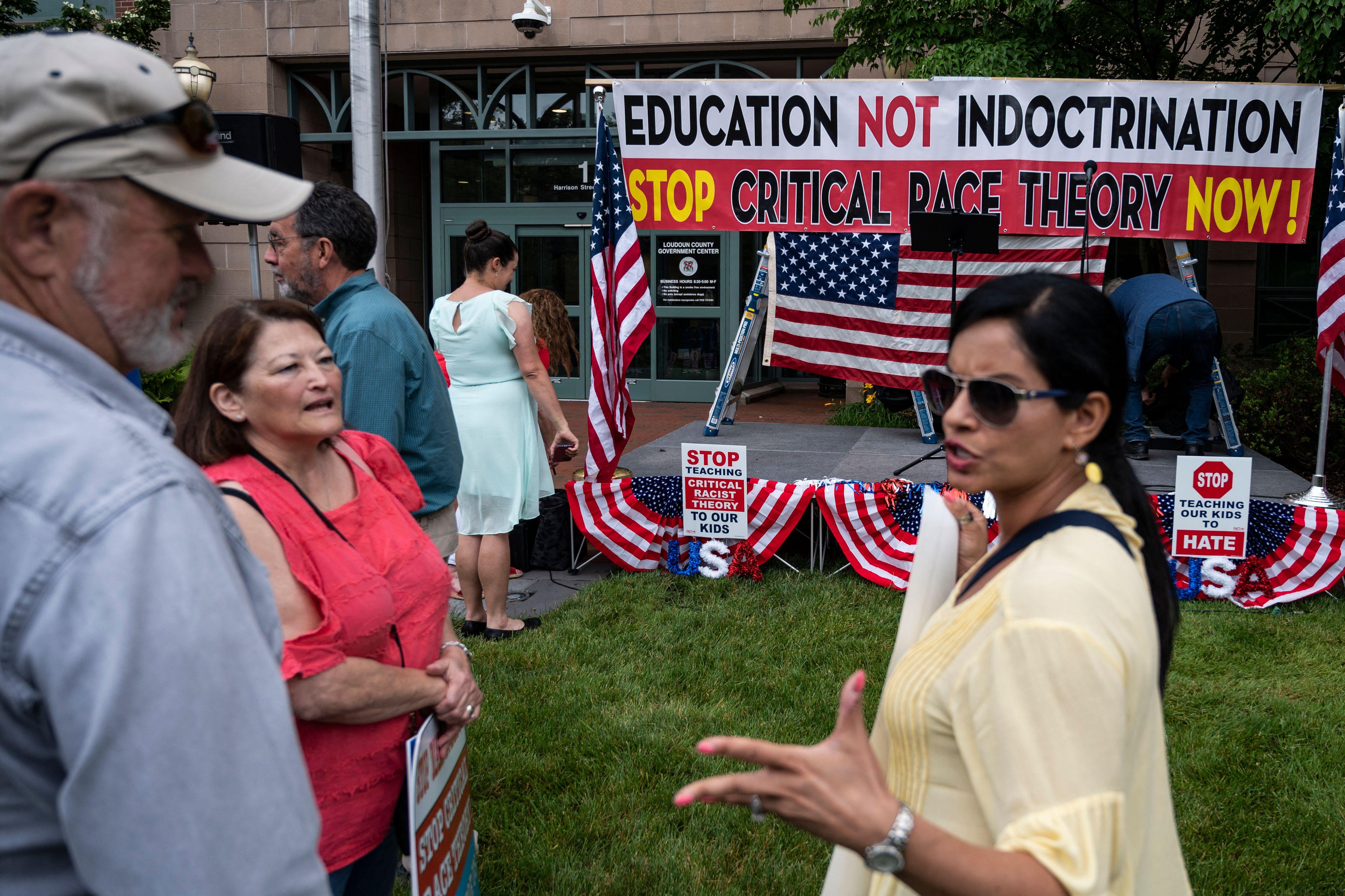 A woman appearing to argue with an older man and woman in front of a sign that says "Education Not Indoctrination, Stop Critical Race Theory Now!"