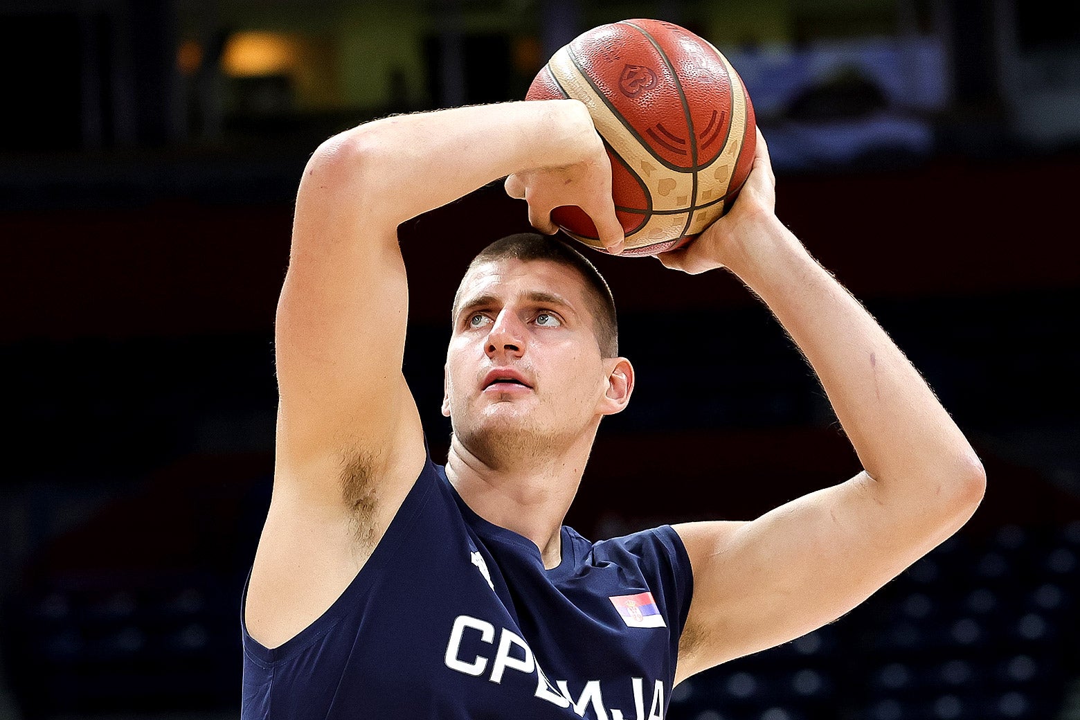Nikola Jokić, wearing a navy jersey, holds a basketball over his head and prepares to throw it.