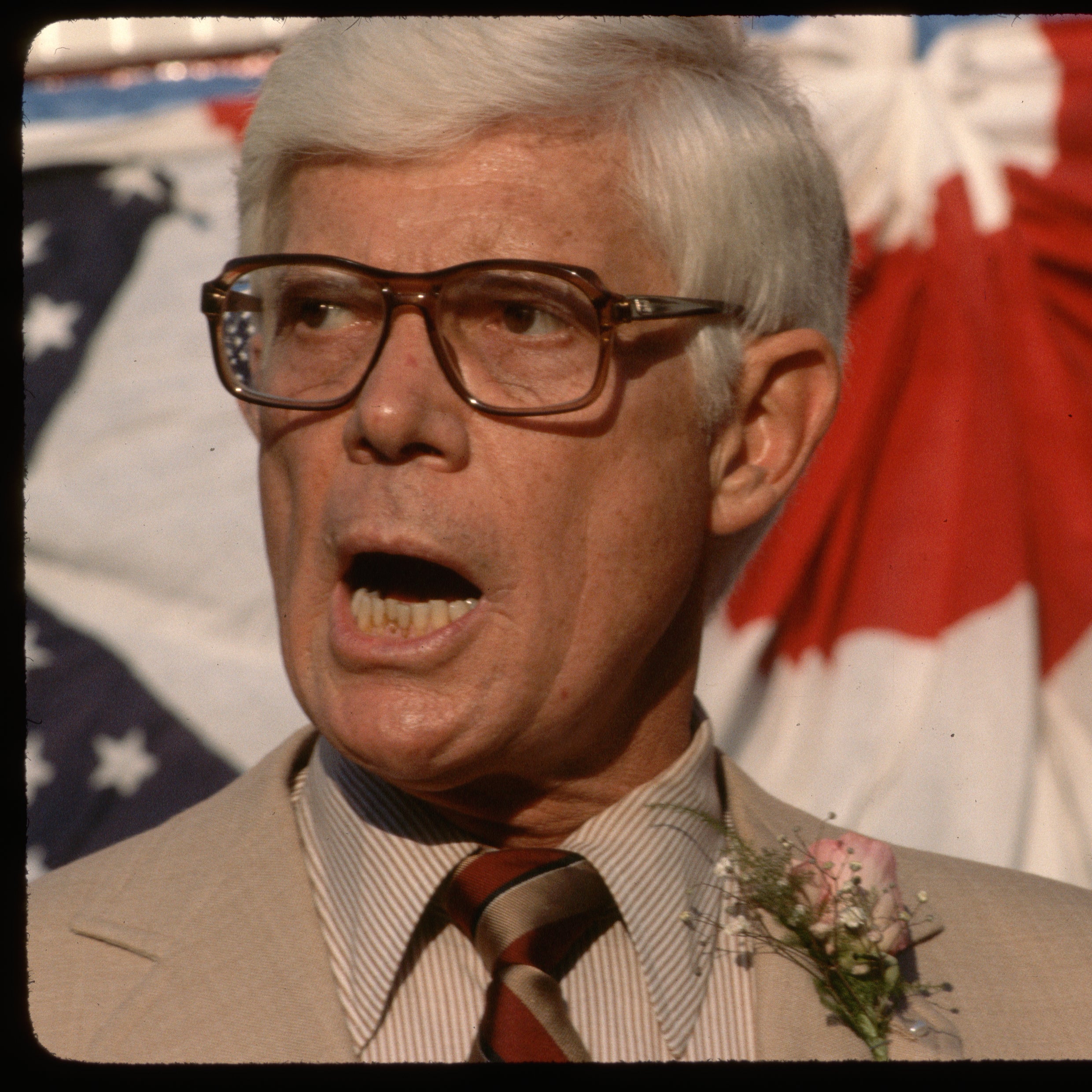 A bespectacled Anderson speaking in front of bunting with a pink boutonniere on his lapel