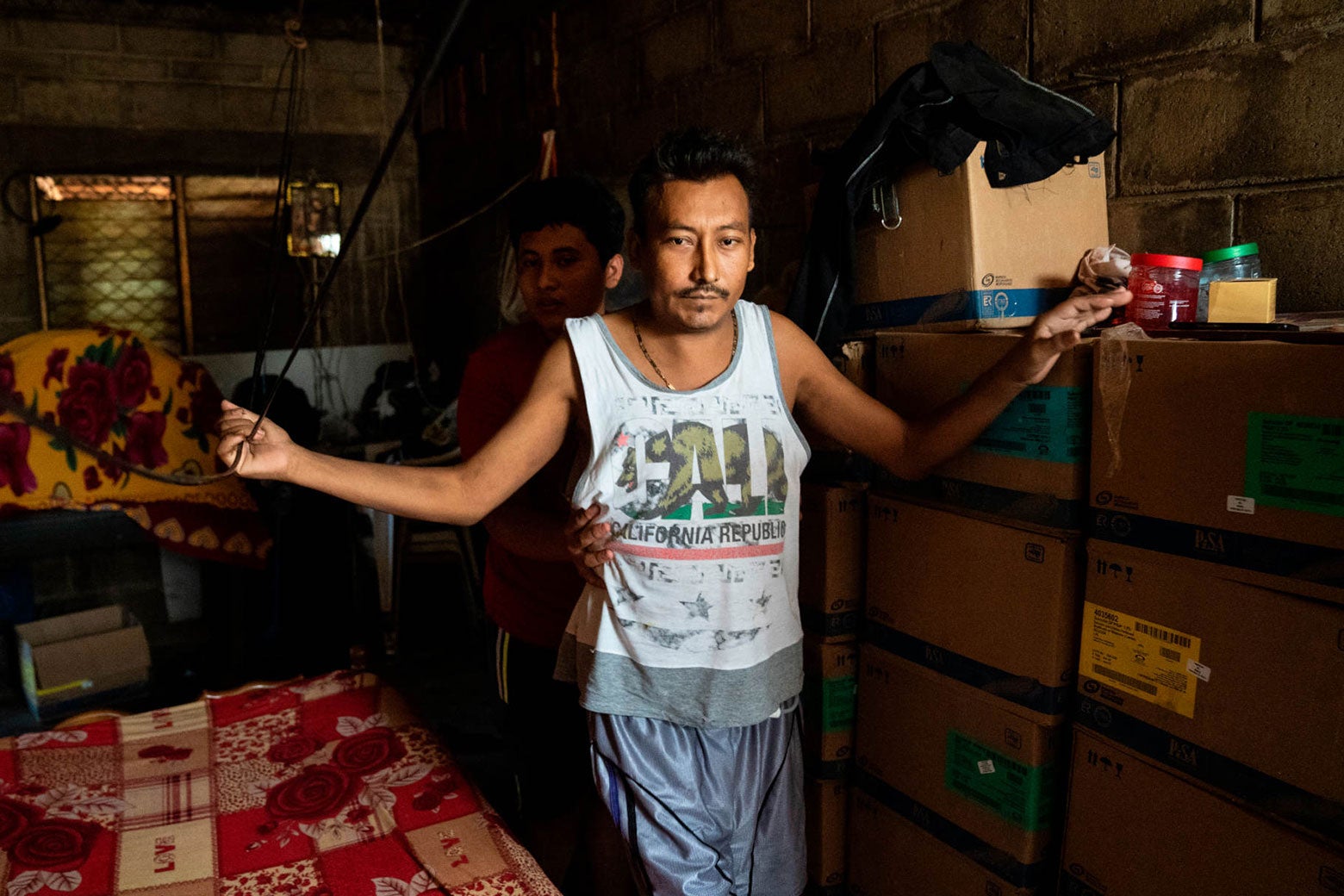 A thin man walks through a house, leaning on a stack of cardboard boxes for stability. A younger boy is behind him, grabbing his waist.