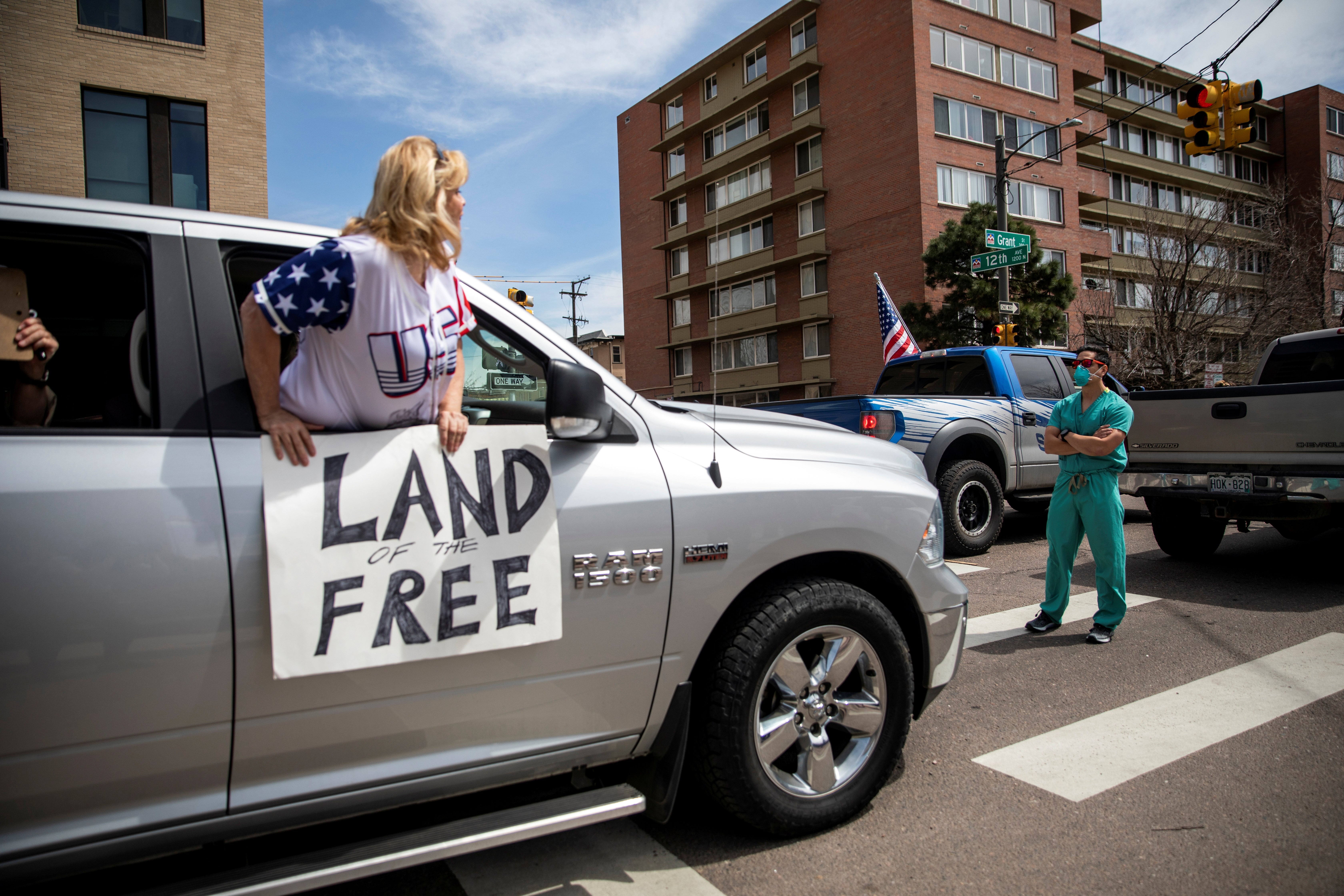 A woman pokes her body out of the passenger seat of a car, carrying a sign saying "Land of the Free." In front of the car, a medical worker in a mask stands with arms crossed.