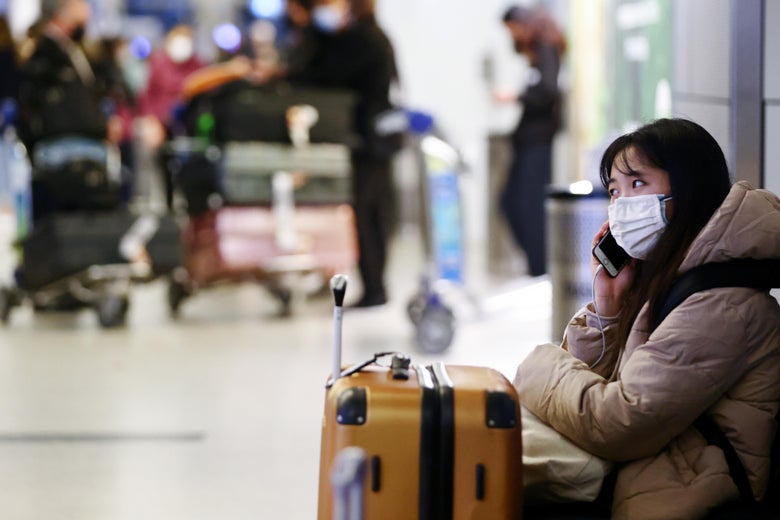 A woman wearing a mask and puffer coat sits next to her luggage as she holds a phone to her ear with other travelers out of focus in the background